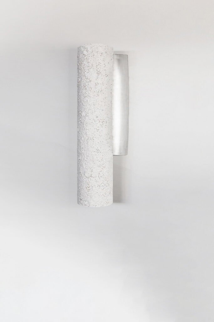 Cylindrical wall light with a rough, stone texture mounted on a plain white wall. The fixture's minimalist design contrasts its textured surface with a smooth, metallic backplate.