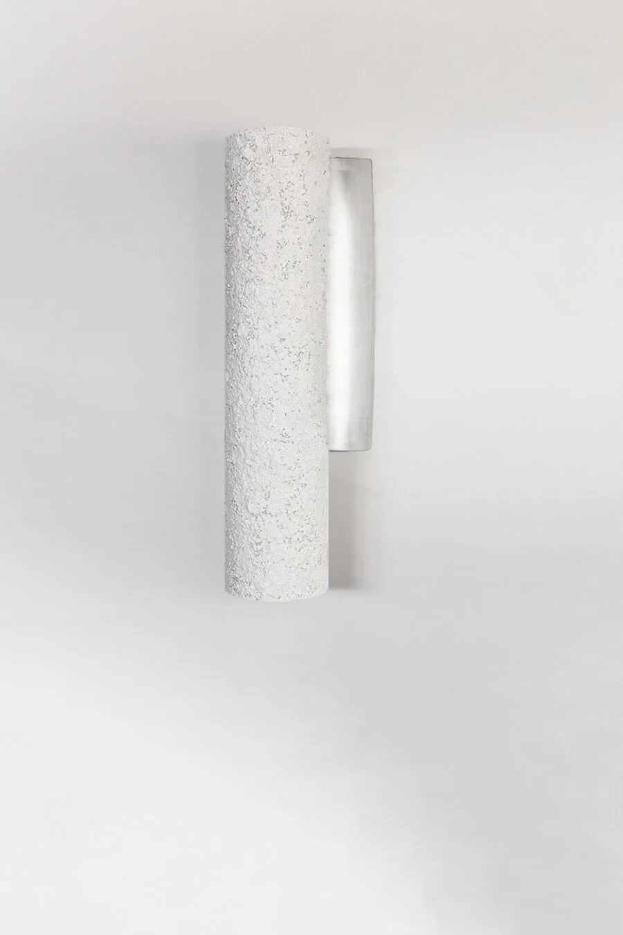 Cylindrical wall light with a rough, stone texture mounted on a plain white wall. The fixture's minimalist design contrasts its textured surface with a smooth, metallic backplate.