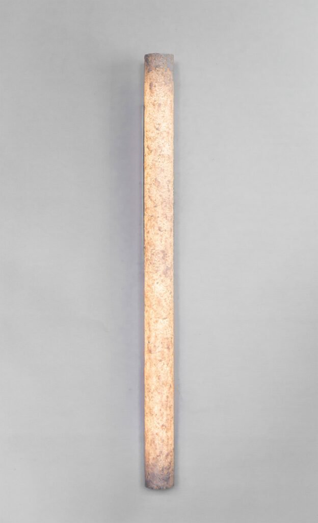 Vertically oriented tube wall sconce with a stone-like texture emitting a warm glow. Its unseen brushed steel mounting rose complements the rustic charm of the textured design with sleek modernity.