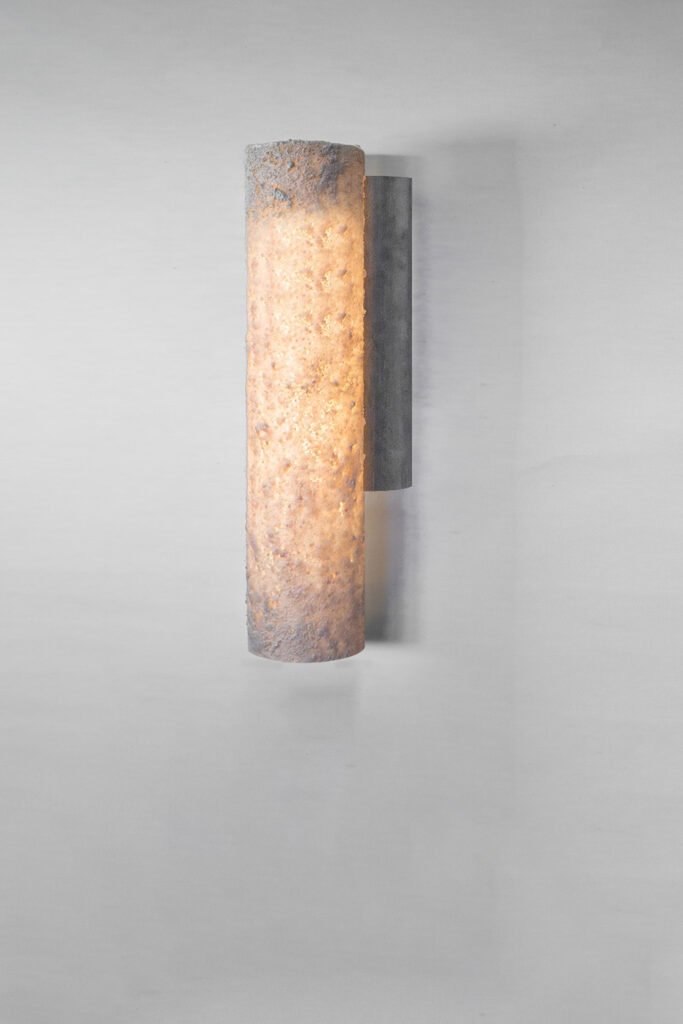 A cylindrical wall sconce with a textured surface emits warm light, mounted on a rectangular backplate against a plain wall. The design blends rugged texture with sleek modern simplicity.