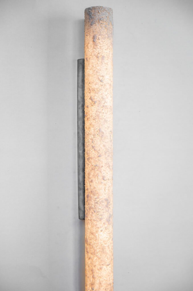 Vertical cylindrical wall sconce with a textured stone-like surface emitting warm, diffused light. Mounted on a brushed steel rose, it blends rugged earthy texture with sleek modern design.