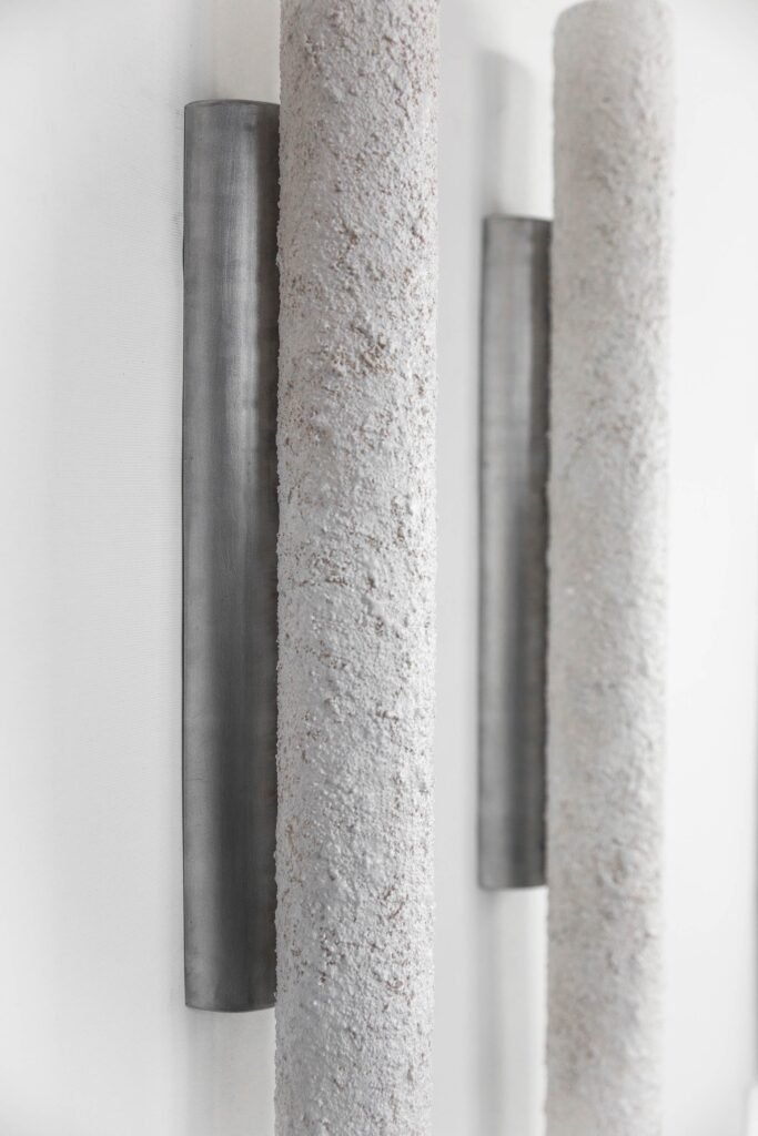 Two vertical cylindrical tube sconces with stone-like textured surfaces on a light-coloured wall. Unlit, they emphasize minimalist design and industrial style against a neutral background.