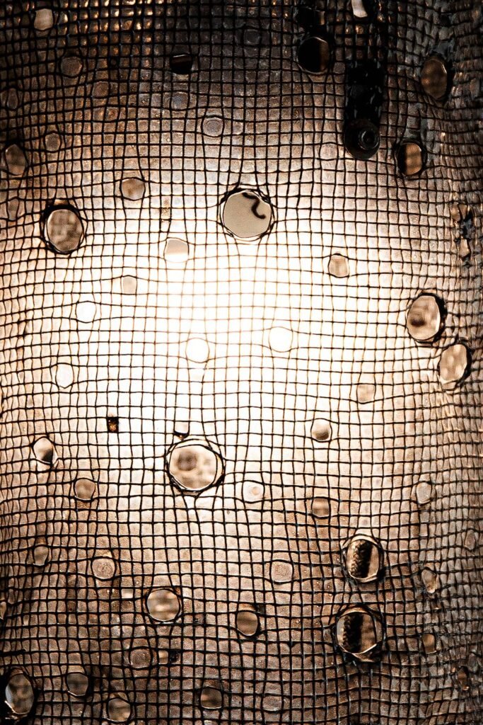 Close-up of a thin, metallic mesh structure with circular holes of varying sizes. Bright light shines through the holes, creating a textured pattern and highlighting the intricate design.