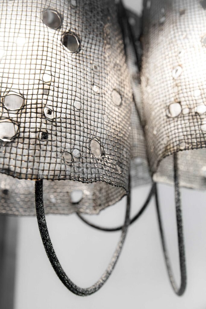 Close-up of cylindrical mesh shades with black polymer finish and circular holes, illuminated to cast intricate patterns. Textile cables hang from the shades shades, enhancing the industrial design.