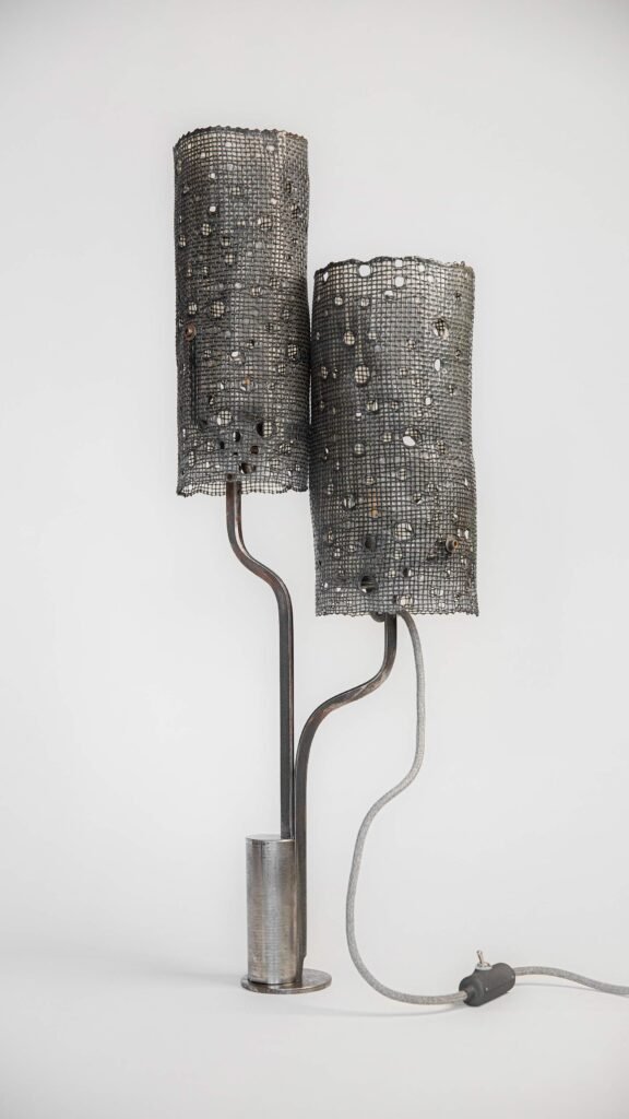 Contemporary lamp with two mesh cylindrical shades featuring circular patterns. Mounted on curved metallic rods with a sleek cylinder base, it has a grey cord and inline switch for industrial charm.
