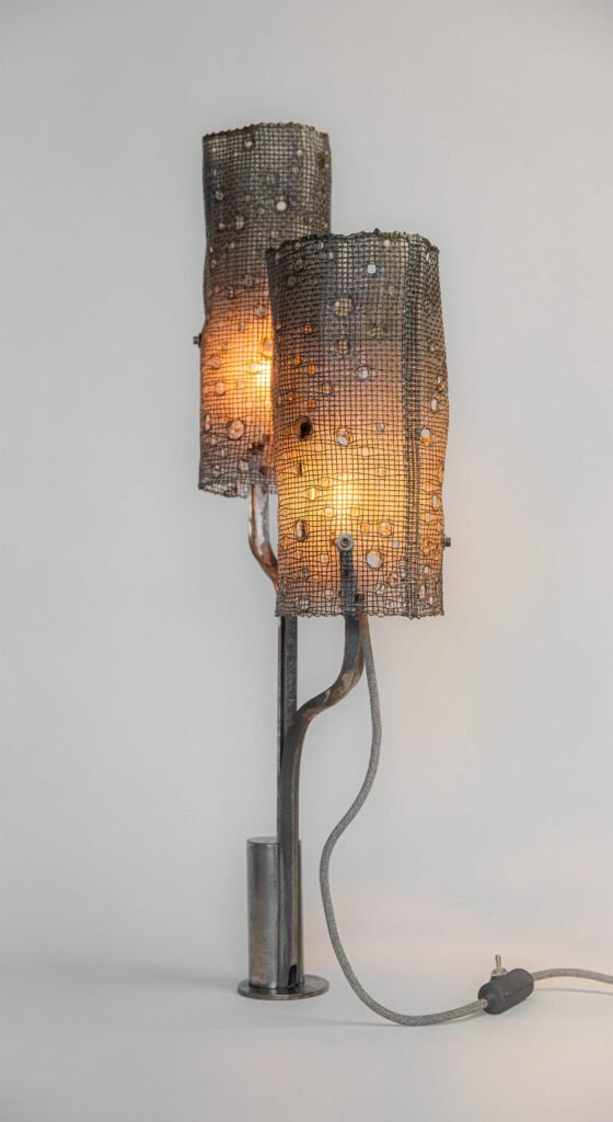 Modern lamp with two illuminated cylindrical mesh shades casting warm light and subtle patterns. Supported by curved steel arms on a round base, it features a grey fabric cord and inline switch.