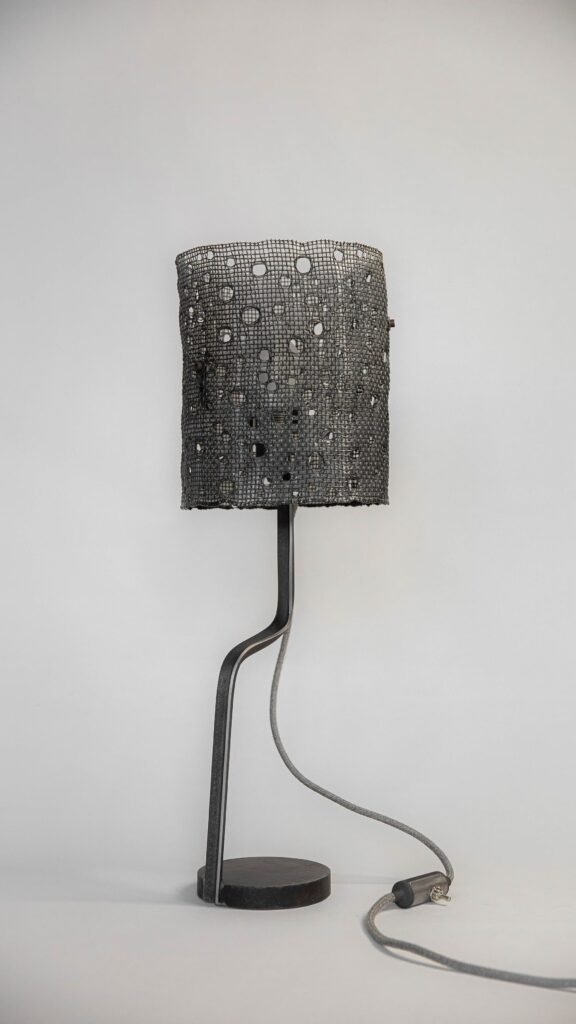 Modern table lamp with an unlit cylindrical mesh shade featuring circular hole patterns. Supported by a curved steel stand and round base, it has a fabric-covered grey cord and minimalist aesthetic.