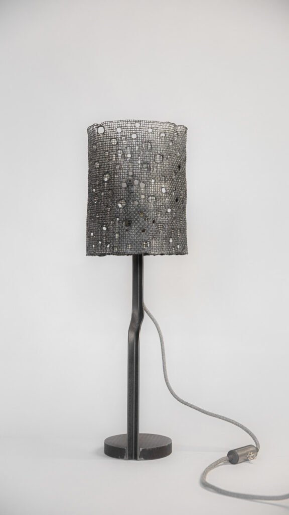 Table lamp with a cylindrical mesh shade featuring circular cut-outs. Supported by a sleek black steel stand, grey fabric cord, and circular base on a white background.