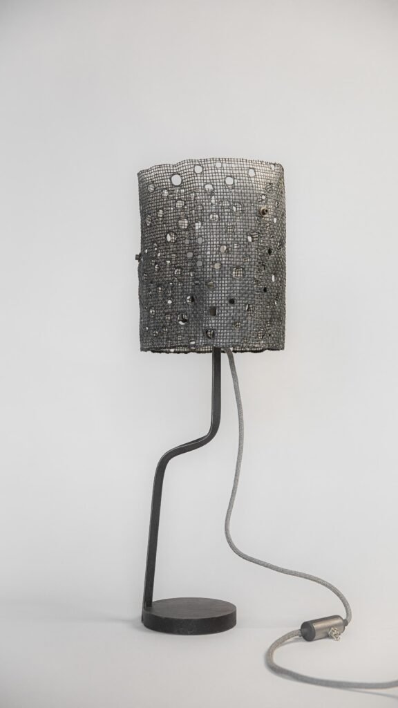 Modern table lamp with an unlit cylindrical mesh shade featuring circular hole patterns. Supported by a curved steel stand and round base, it includes a fabric-covered grey cord and inline switch.