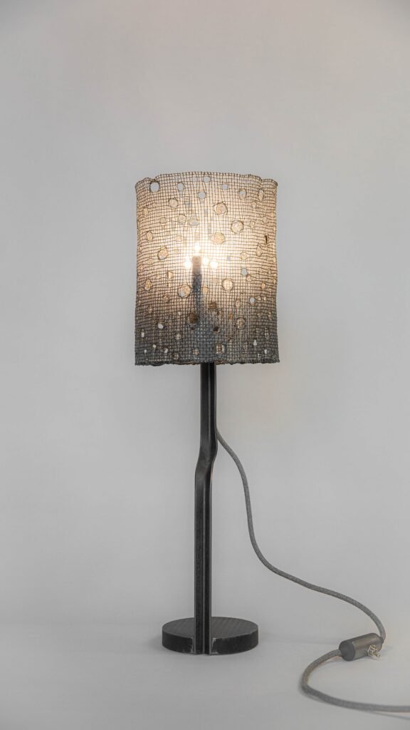 Modern table lamp with a mesh cylindrical shade featuring circular holes and a gradient to black. Supported by a curved steel stand and round base, it includes an outward-extending textile cord.