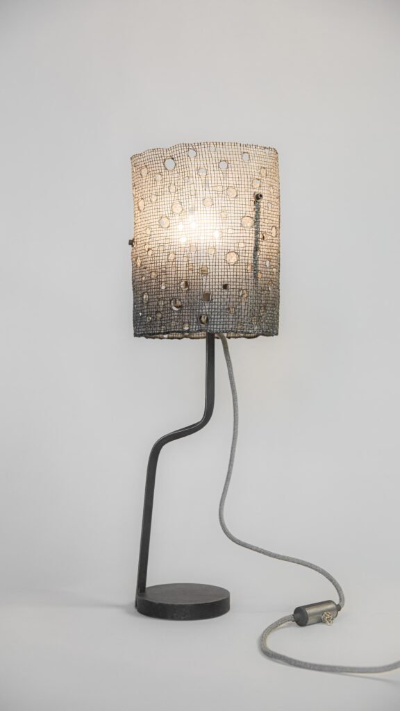 Modern lamp with illuminated cylindrical mesh shade casting intricate patterns. Curved steel stand on round base, with grey fabric-covered cord featuring an inline switch. Neutral backdrop.
