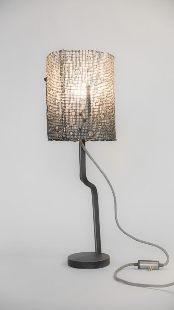Modern table lamp with lit cylindrical mesh shade featuring circular hole patterns. Supported by curved steel stand and round base, with grey fabric cord and inline switch against neutral backdrop.