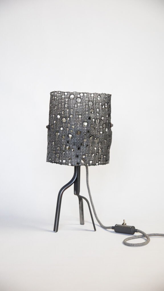 Unlit minimalist lamp with steel base, curved legs, cylindrical mesh shade with organic pattern and bolts. Fabric-covered cord with toggle switch adds refinement, enhancing its industrial aesthetic.