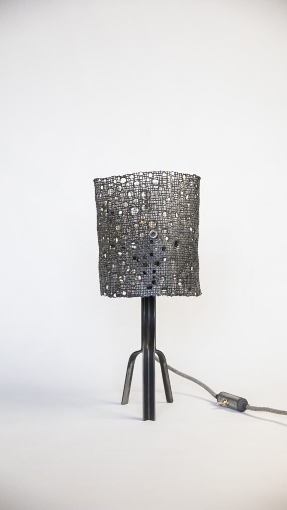 Unique lamp with black mesh cylindrical shade featuring circular holes, sleek steel tripod stand. Includes a grey cylindrical switch on the textile power cord, highlighted against a white backdrop.