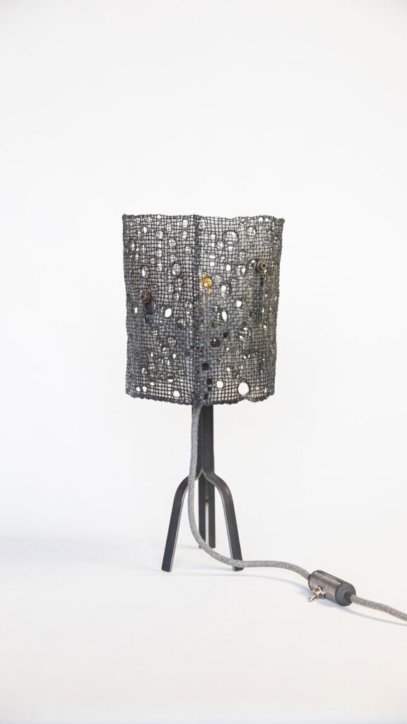Stylish lamp with black cylindrical mesh shade featuring circular hole patterns on a sleek tripod stand with three thin legs. Fabric grey power cord with inline switch, set against a white backdrop.