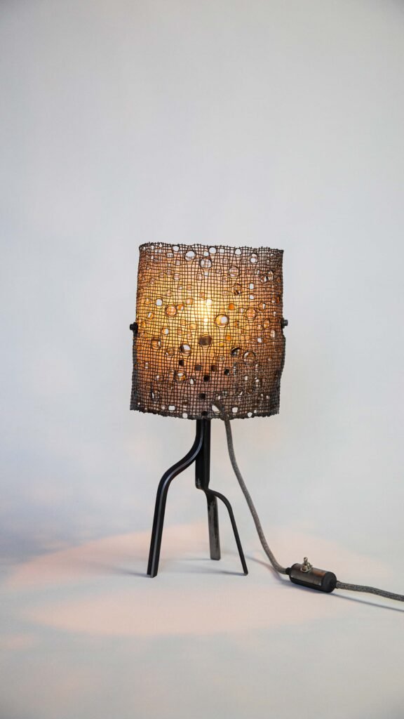Table lamp with a steel mesh cylindrical shade featuring circular holes, casting captivating patterns. Tripod stand with three slender legs, fabric cable with inline switch on a light background.