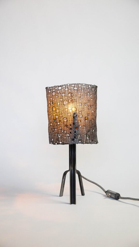Modern lamp with dark steel tripod base and perforated mesh shade coated in polymer. Organic patterns diffuse warm light, casting intricate shadows. Includes fabric-covered cord with inline switch.