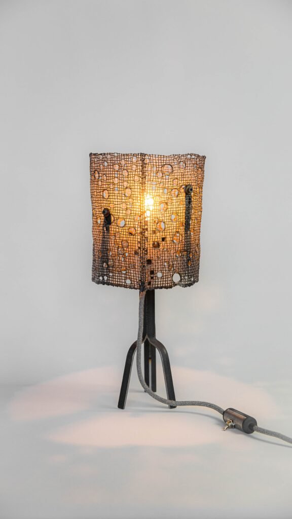 Contemporary lamp with dark steel tripod base and cylindrical polymer-coated mesh shade featuring organic perforations, fabric-covered cord for an industrial look. Exposed bulb casts warm patterns,