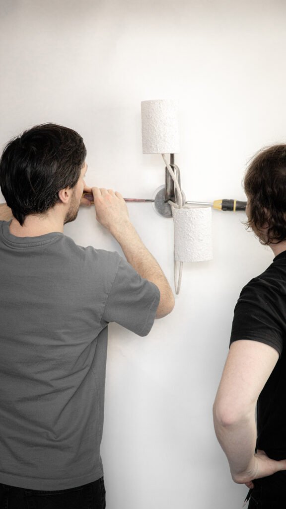 Two people install a wall sconce with textured stone-effect cylindrical shades on curved steel arms. Textile cables and lights off highlight design and functionality during fitting.