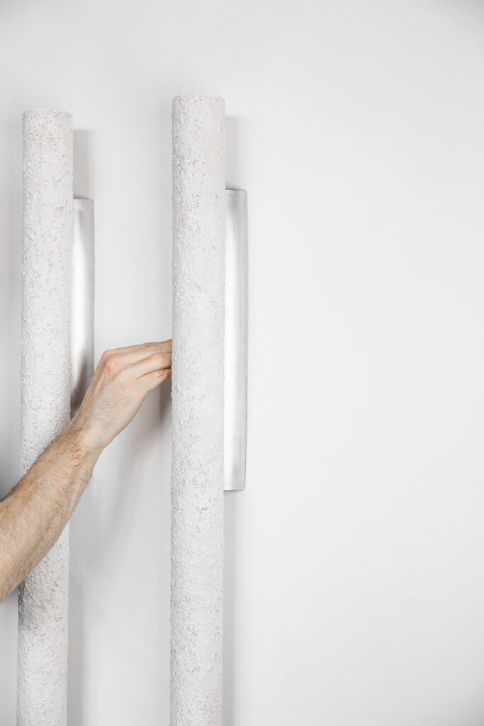 Two unlit vertical tube wall sconces on a light-coloured wall. Textured stone-like surfaces and brushed steel roses add sleek contrast, with a hand interacting with one fixture.