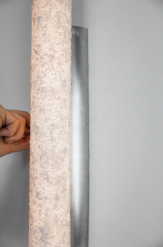 Close-up of a hand touching a cylindrical tube sconce with a textured stone-like surface. Rugged texture contrasts with the smooth brushed steel rose, while soft light enhances the modern design.