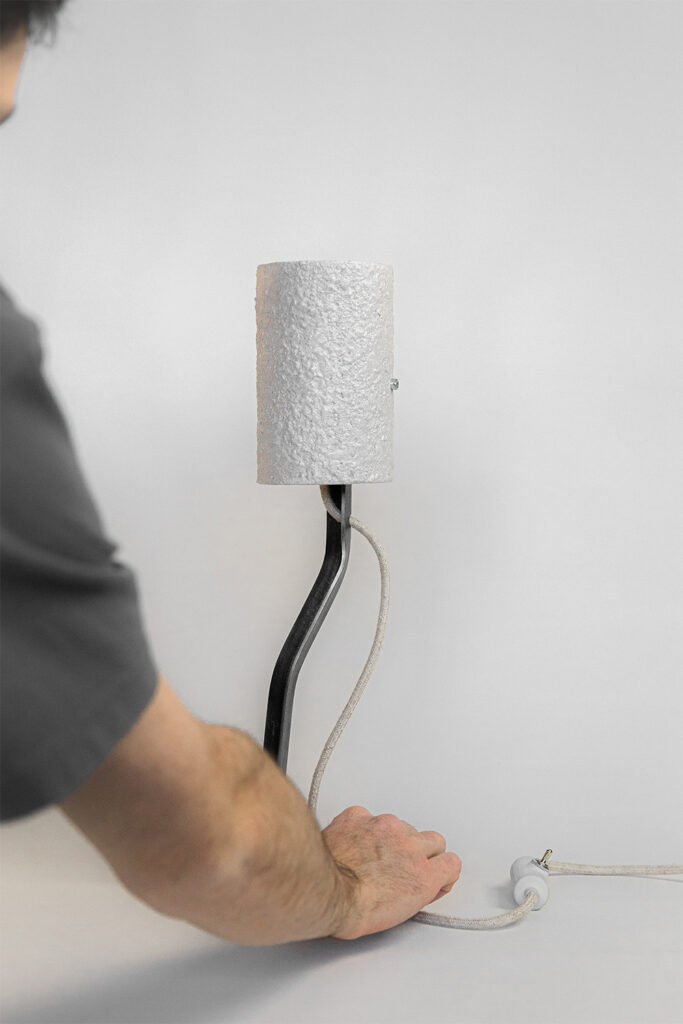 Modern table lamp with a curved metal stand, neutral-tone textured shade, and fabric-covered cord. Person adjusts its position, showcasing its minimalist, industrial-organic design.