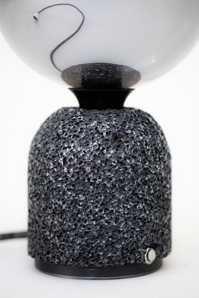 Modern table lamp with a porous dark aluminum foam base and a mirrored globe bulb. A textile-covered power cable extends from the base, with only a short section visible directly, while the full length of the cable appears clearly in the bulb’s reflection