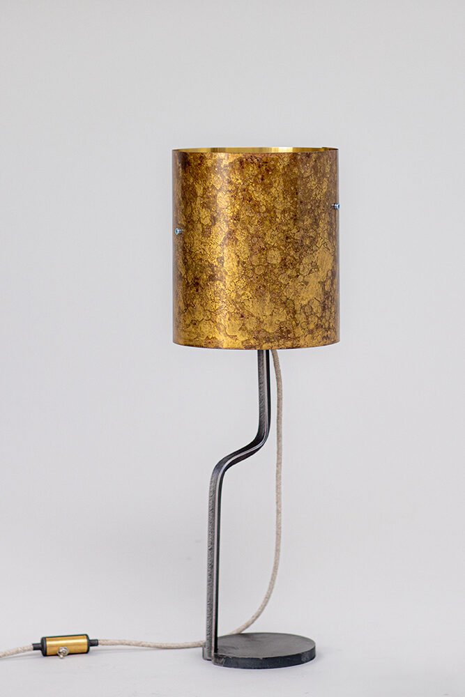 Modern lamp with marbled brass patina cylindrical shade, curved steel stand, round base, and fabric-covered cord with brass inline switch. Presented against a plain white backdrop for emphasis.