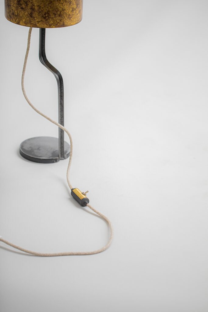 Lamp with cylindrical mesh shade featuring a textured pattern in brownish-yellow tones. Supported by curved metal stand and round base. A beige cord with black-and-gold switch lies on a light surface.