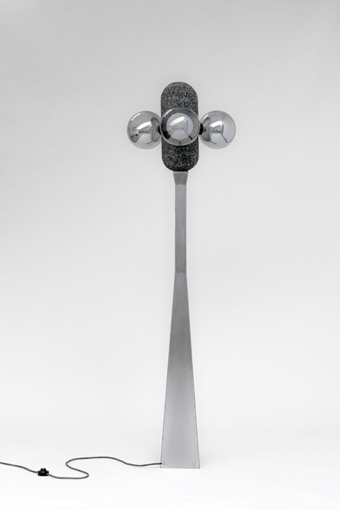 Contemporary floor lamp with a tapered steel base, aluminium foam texture, and symmetric metallic spheres. A power cord hints at its modern, functional design.