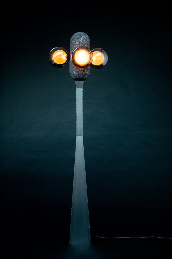 Modern floor lamp with a sleek steel base, aluminium foam top, and three spherical bulbs glowing orange. A sculptural design set against a dark backdrop.
