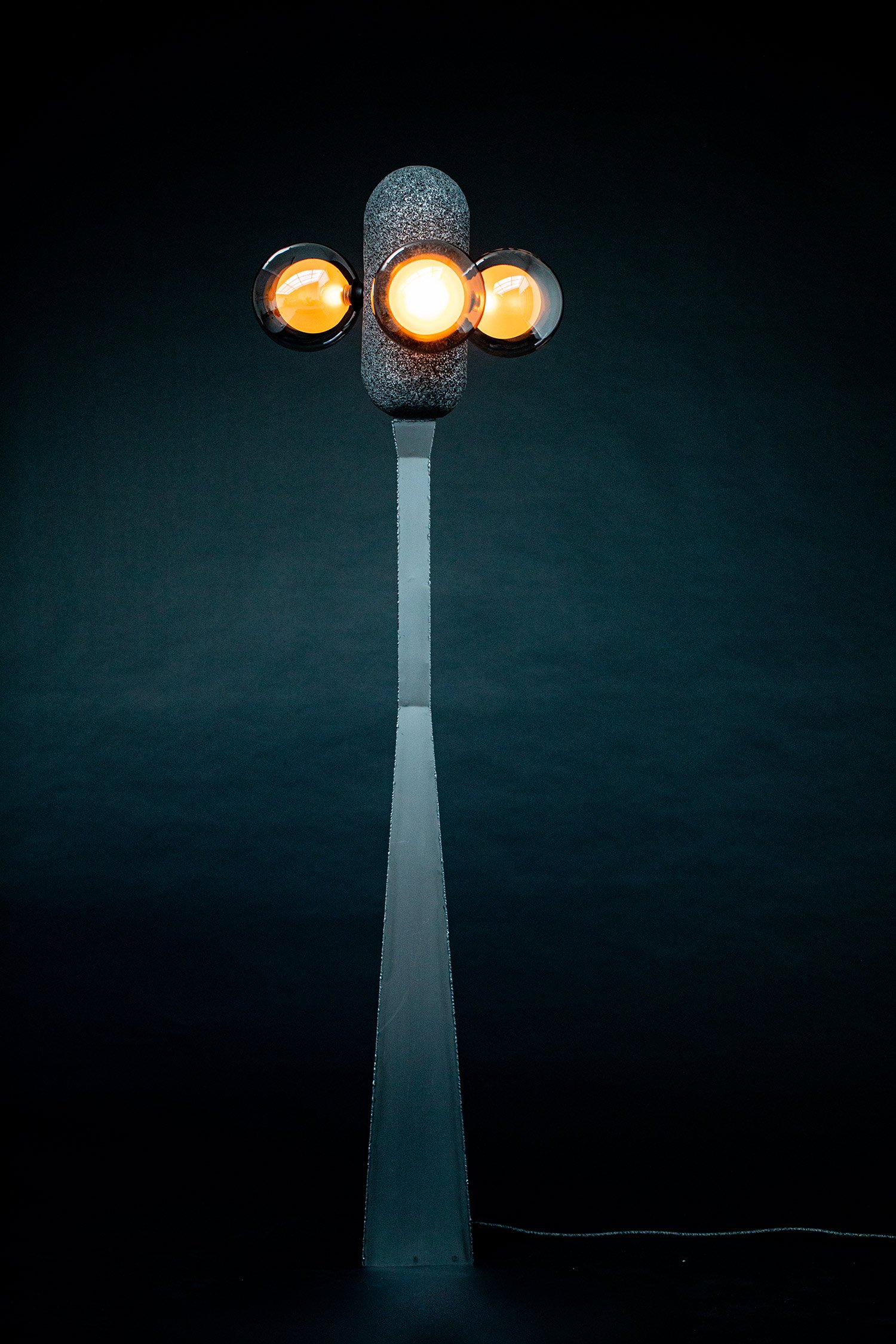 Modern floor lamp with a sleek steel base, aluminium foam top, and three spherical bulbs glowing orange. A sculptural design set against a dark backdrop.