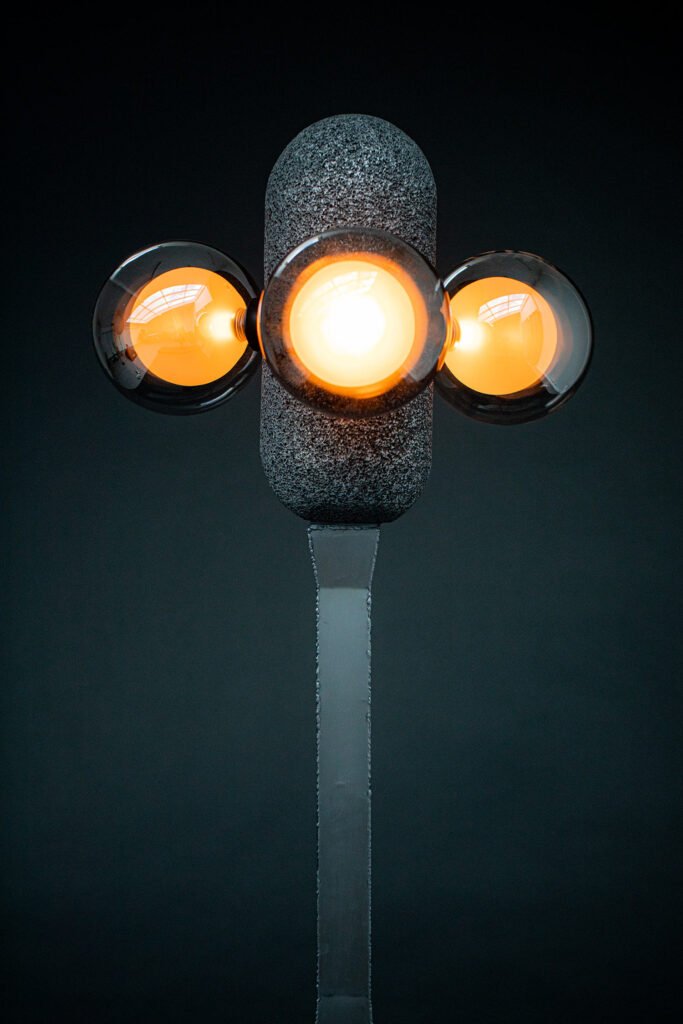 Modern floor lamp with a steel base and aluminium foam top. Three glowing orange spherical bulbs create warm light, blending industrial materials with sculptural elegance