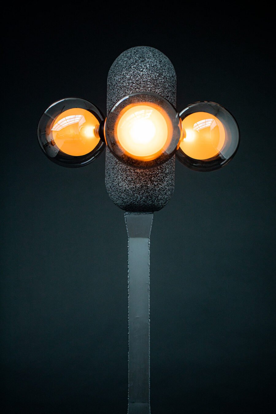 Modern floor lamp with a steel base and aluminium foam top. Three glowing orange spherical bulbs create warm light, blending industrial materials with sculptural elegance