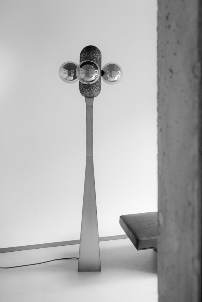 Modern floor lamp with a tapered steel base, aluminium foam top, and three horizontal spherical bulbs emitting soft light. Set against a minimalist backdrop.
