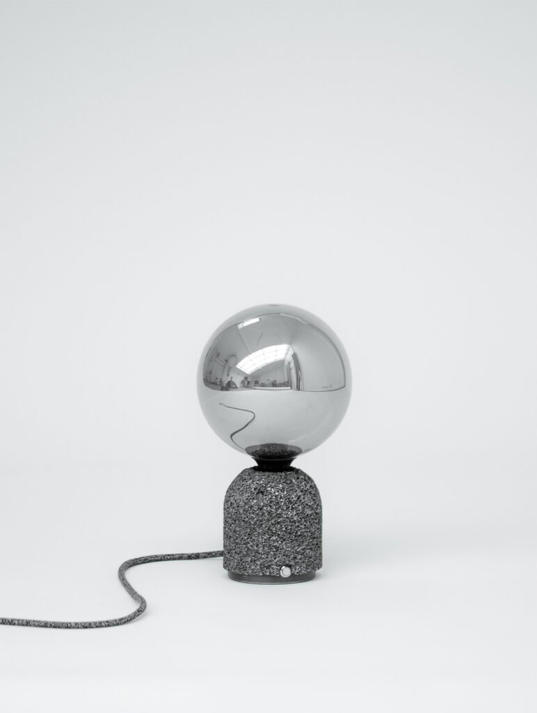 Modern table lamp with a dark aluminum foam base and a mirrored globe bulb, shown against a plain white background with its fabric-covered power cord extending outward.