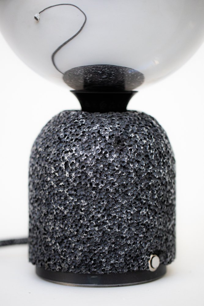Modern table lamp with a porous dark aluminum foam base and a mirrored globe bulb. A textile-covered power cable extends from the base, with only a short section visible directly, while the full length of the cable appears clearly in the bulb’s reflection