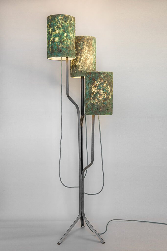 Modern floor lamp with three patina-textured cylindrical shades emitting warm, diffused light. Shades at varied heights on black angular frame with tripod base. Visible cords on a neutral wall.