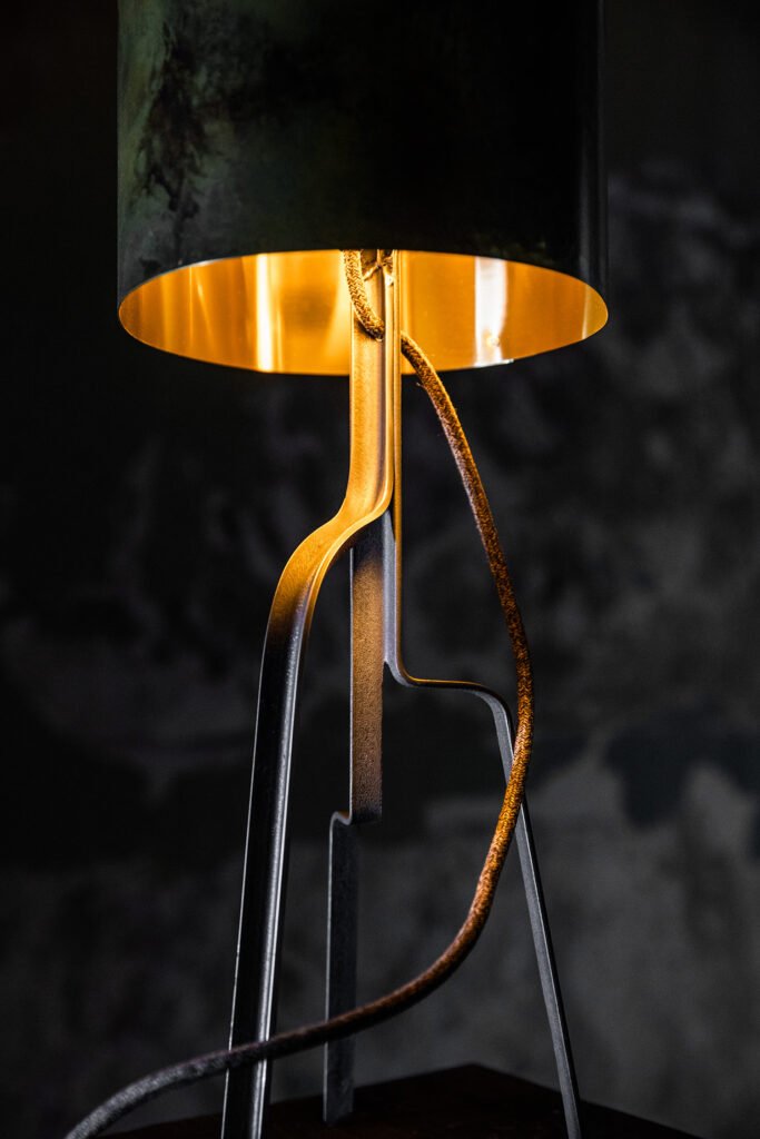 Modern-industrial lamp with weathered cylindrical steel shade, gold interior, and curved grey steel base. Fabric power cord with brass switch complements its warm glow and aged, moody backdrop.